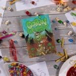 Goblin Quest front cover on a white wooden table surrounded by pens, paper and snacks - © SHAW STUDIO