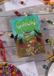Goblin Quest front cover on a white wooden table surrounded by pens, paper and snacks - © SHAW STUDIO