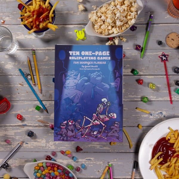 Front cover of Ten One-Page RPGs for Younger Players on a white table surrounded by dice, pencils and snacks © SHAW STUDIO